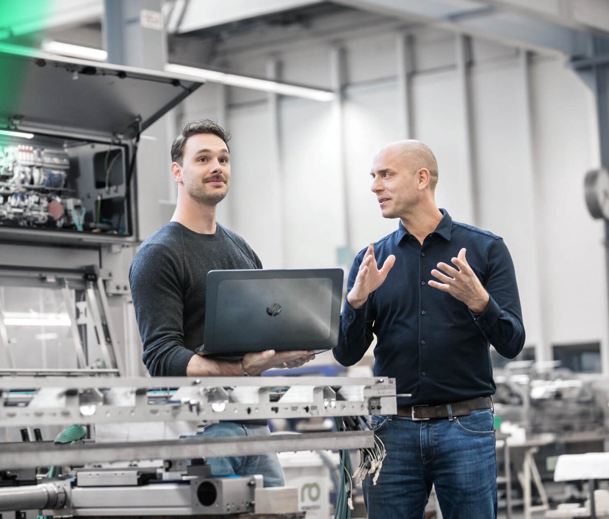 Two engineers discussing in front of industrial machinery, one holding a laptop.
