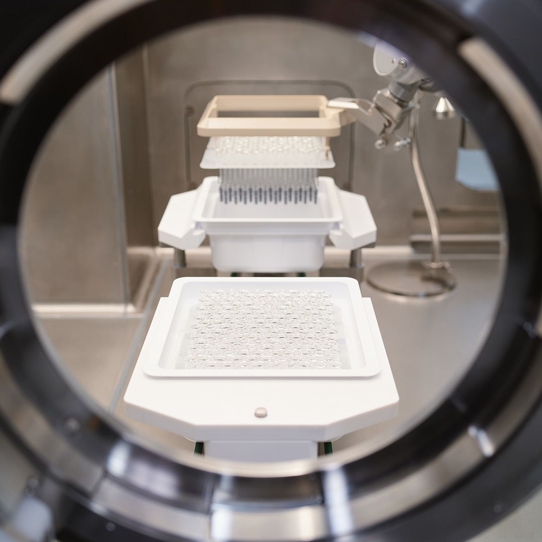 View through a round isolator window onto several white trays loaded with ready to use containers inside an aseptic filling cell.
